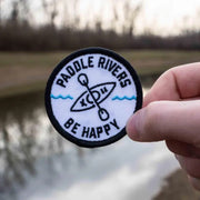 A hand holding an embroidered patch with the text 'Paddle Rivers - BE HAPPY KAYAKING' and an illustration of a kayak on the patch.