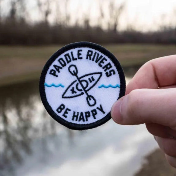 A hand holding an embroidered patch with the text 'Paddle Rivers - BE HAPPY KAYAKING' and an illustration of a kayak on the patch.