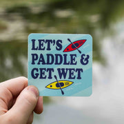 A hand holding a rectangular sticker with the text 'LET'S PADDLE & GET WET' and illustrations of a paddle and water.