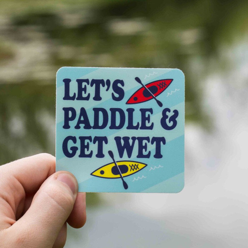 A hand holding a rectangular sticker with the text 'LET'S PADDLE & GET WET' and illustrations of a paddle and water.
