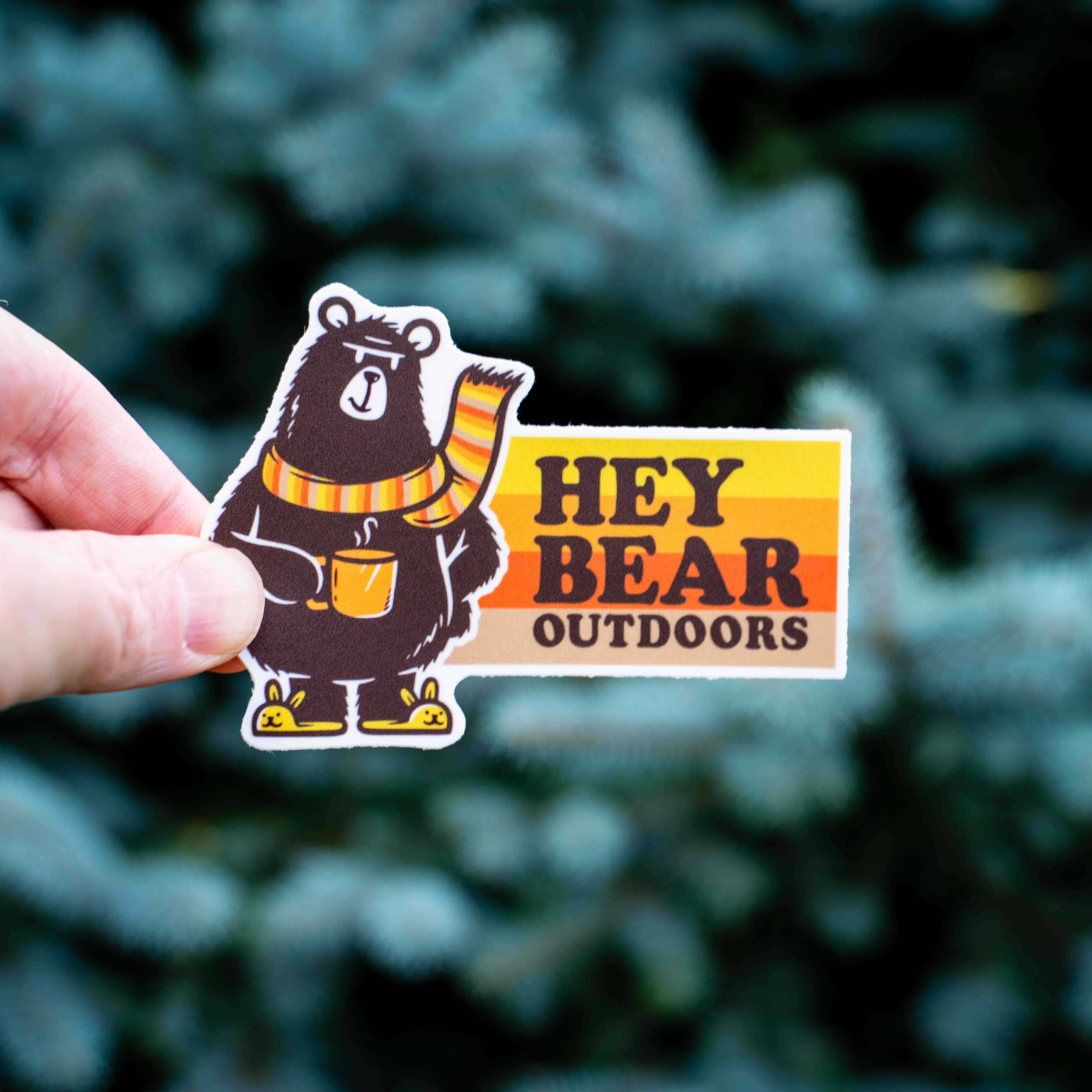 Hand holding a 'Hey Bear Outdoors' sticker with a bear graphic against a blurred green background.