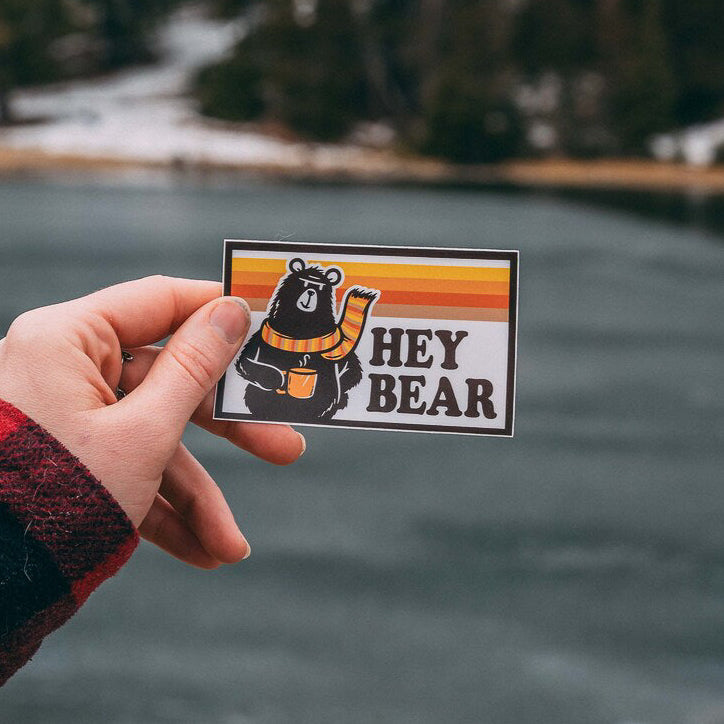 A sticker with a bear design and the text 'Hey Bear' held against a backdrop of a snowy landscape with trees.