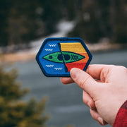 A hand holding a square-shaped embroidered patch with a kayak and fish design, against a blurred natural background.