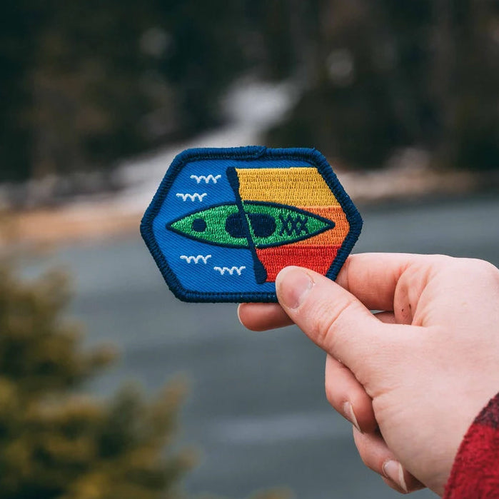 A hand holding a square-shaped embroidered patch with a kayak and fish design, against a blurred natural background.