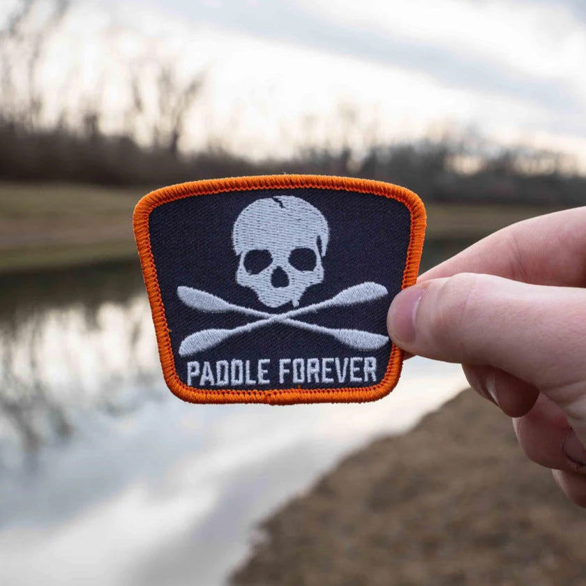 A hand holding an embroidered patch with the text 'Paddle Forever' and an image of a skull and crossed bones, against a backdrop of a body of water and trees.