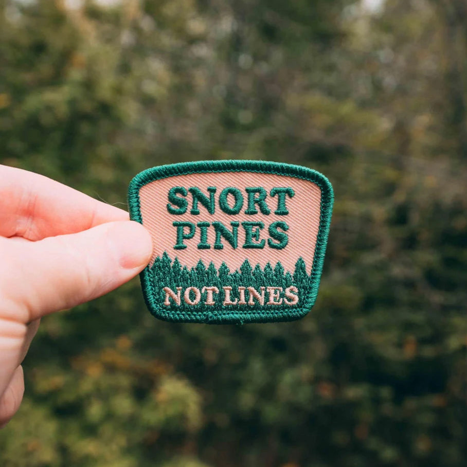 A hand holding an embroidered patch with the text 'Snort Pines Not Lines' and a pine tree logo.